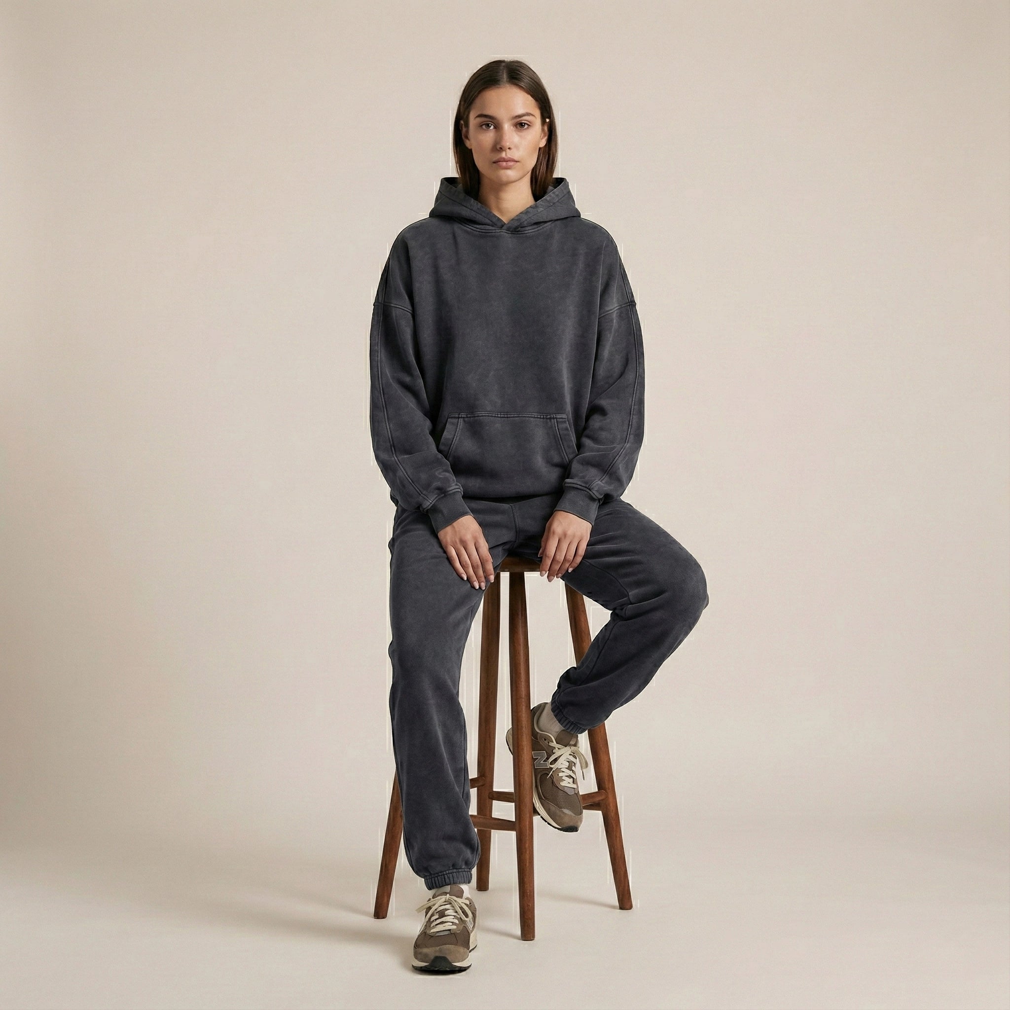 Person wearing a dark gray hoodie and pants sitting on a wooden stool against a beige background