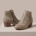 A pair of taupe faux-leather ankle boots with geometric laser-cut side panels and a stacked block heel, displayed on a light beige platform against a neutral background.