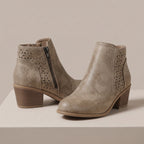 A pair of taupe faux-leather ankle boots with geometric laser-cut side panels and a stacked block heel, displayed on a light beige platform against a neutral background.
