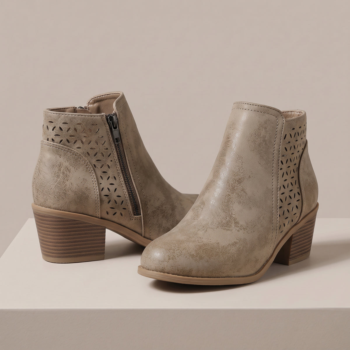 A pair of taupe faux-leather ankle boots with geometric laser-cut side panels and a stacked block heel, displayed on a light beige platform against a neutral background.