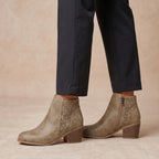 Taupe suede ankle boots with geometric laser-cut side panels and a low block heel, shown on a model wearing dark trousers against a warm beige background.