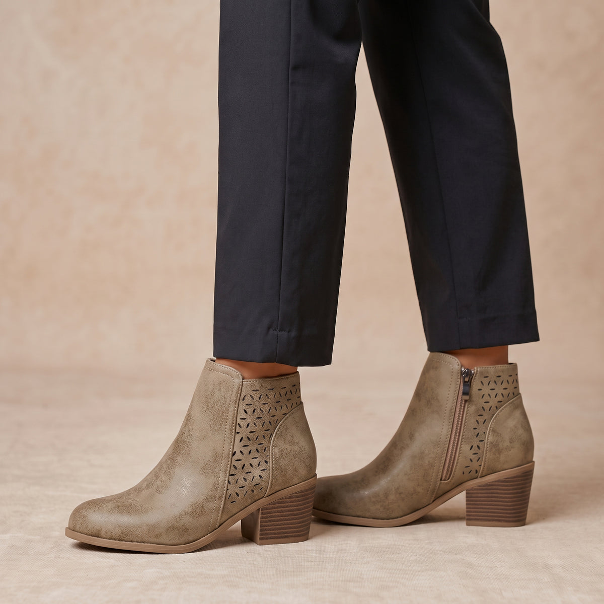 Taupe suede ankle boots with geometric laser-cut side panels and a low block heel, shown on a model wearing dark trousers against a warm beige background.