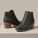 A pair of dark gray faux-leather ankle boots with geometric laser-cut side panels and stacked block heels, displayed on a light beige platform against a neutral beige background.