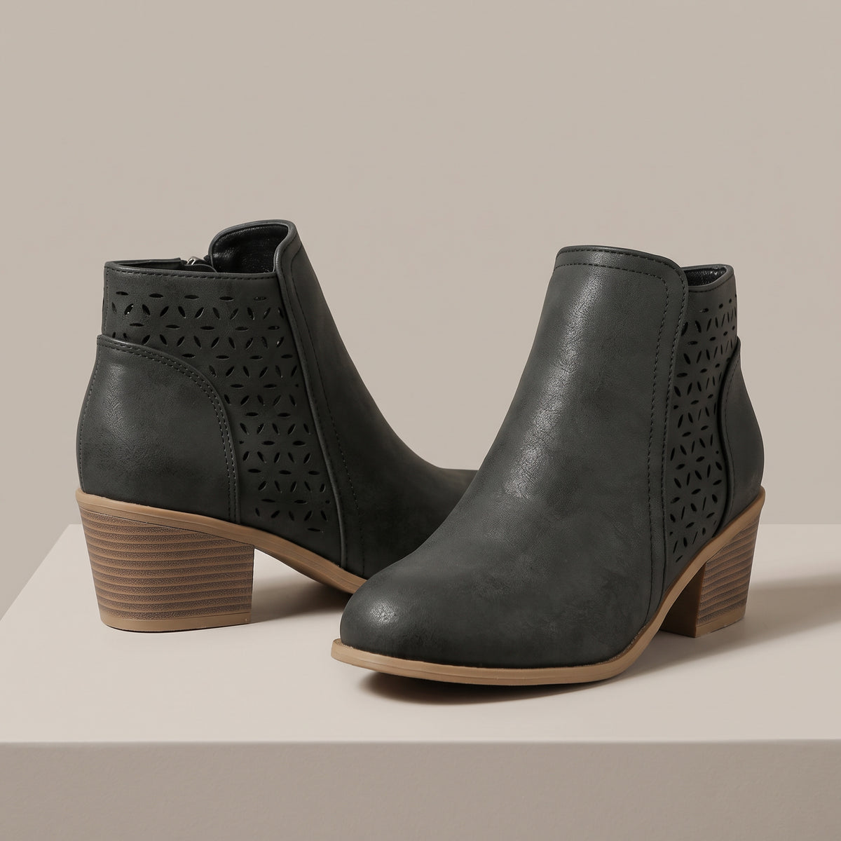 A pair of dark gray faux-leather ankle boots with geometric laser-cut side panels and stacked block heels, displayed on a light beige platform against a neutral beige background.
