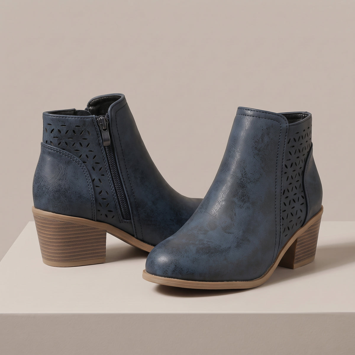 A pair of blue faux-leather ankle boots with laser-cut geometric patterns on the sides, a side zipper, and a stacked block heel, displayed on a light beige platform against a neutral beige background.