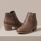 A pair of brown faux-leather ankle boots with distressed textures, geometric laser-cut side panels, and stacked block heels, displayed on a light beige platform against a neutral beige background.