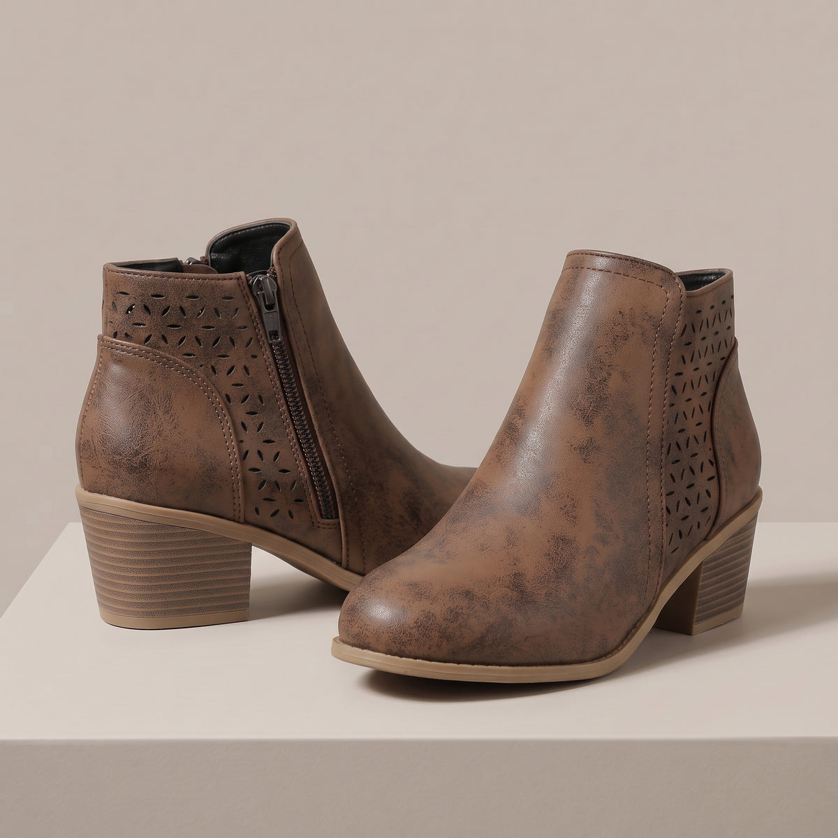 A pair of brown faux-leather ankle boots with distressed textures, geometric laser-cut side panels, and stacked block heels, displayed on a light beige platform against a neutral beige background.