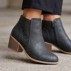 A pair of women's dark gray faux-leather ankle boots with laser-cut geometric patterns on the sides, a low block heel, and a tan sole, shown on a model wearing dark trousers.