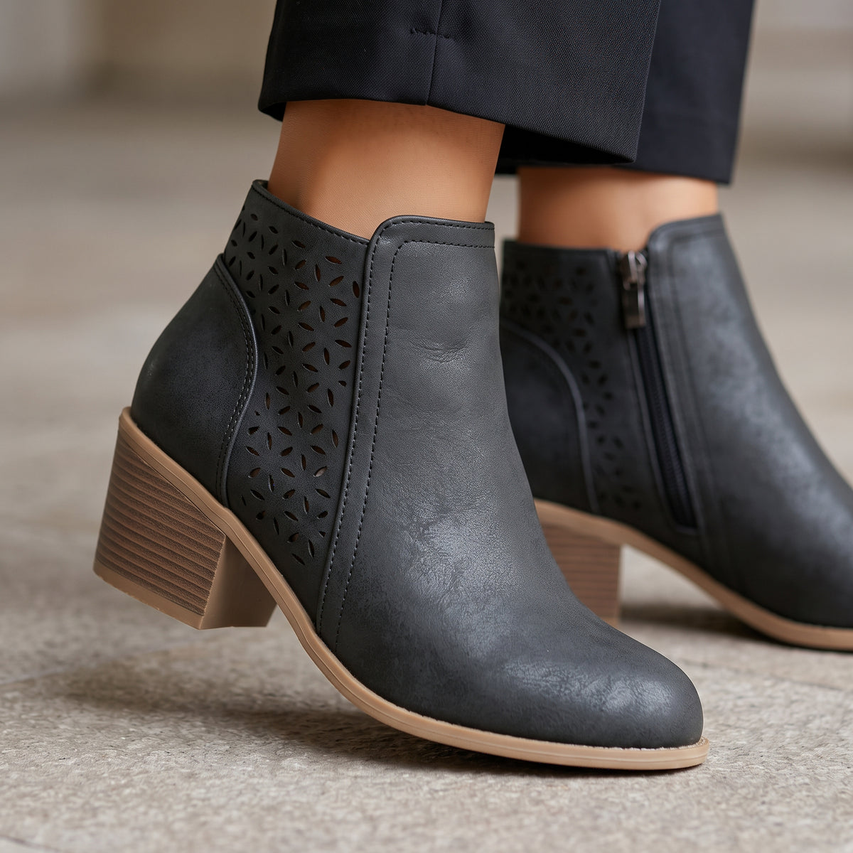 A pair of women's dark gray faux-leather ankle boots with laser-cut geometric patterns on the sides, a low block heel, and a tan sole, shown on a model wearing dark trousers.