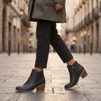 A person walking on a city street wearing black ankle boots with side cutouts and stacked block heels, paired with dark trousers and a gray coat.