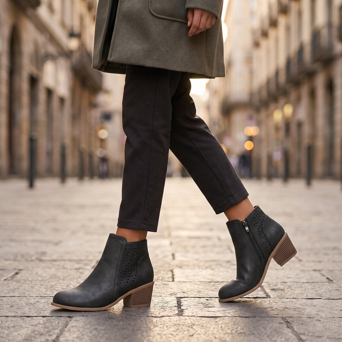 A person walking on a city street wearing black ankle boots with side cutouts and stacked block heels, paired with dark trousers and a gray coat.