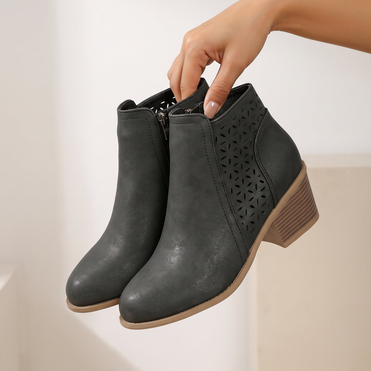 A pair of dark gray ankle boots with laser-cut geometric patterns on the sides and a low block heel, held by a hand against a neutral beige background.