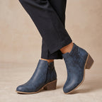 A pair of women's blue leather ankle boots with laser-cut geometric patterns on the sides, shown on a model wearing dark trousers against a warm beige background.