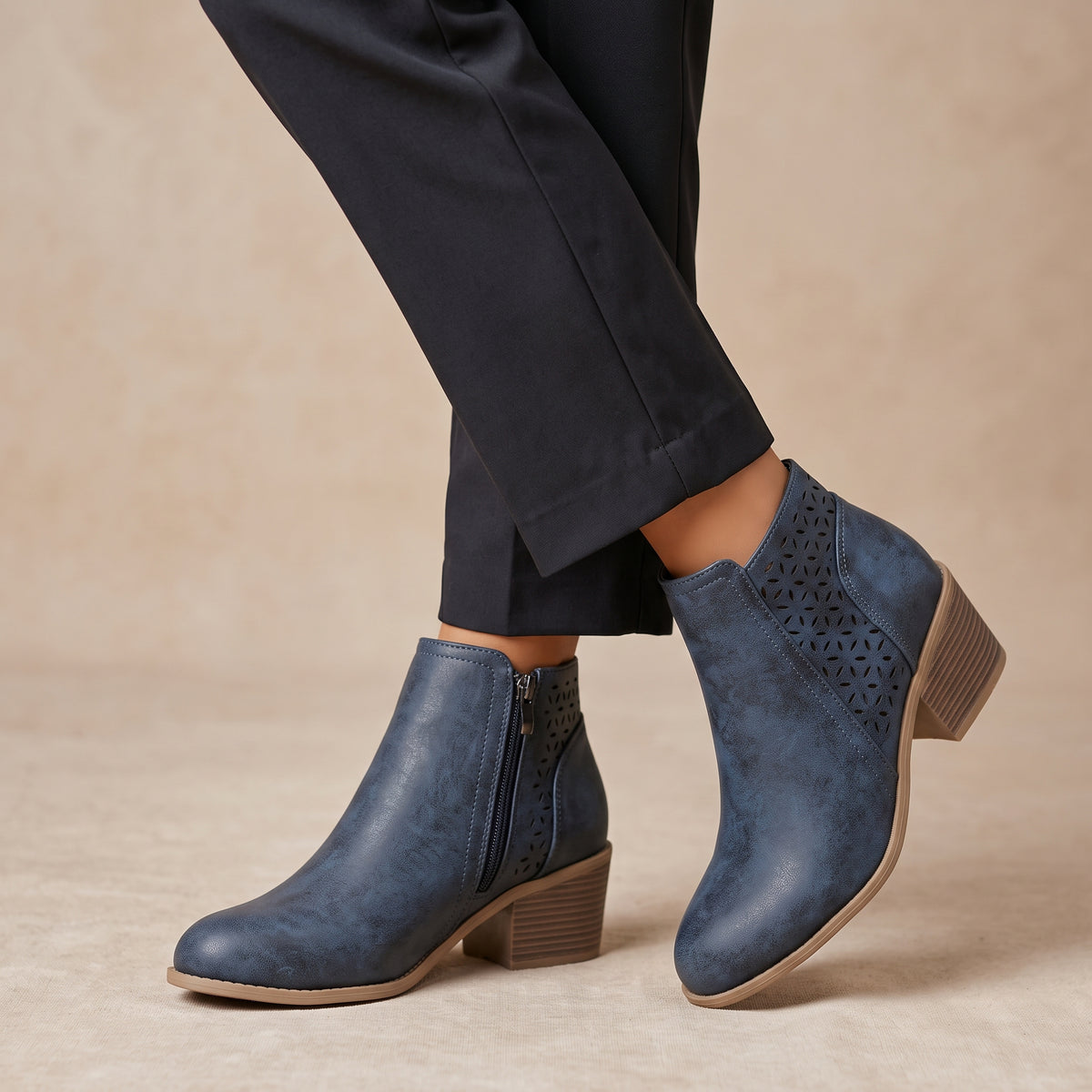 A pair of women's blue leather ankle boots with laser-cut geometric patterns on the sides, shown on a model wearing dark trousers against a warm beige background.