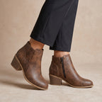 A pair of women's brown ankle boots with a distressed finish, laser-cut geometric pattern on the side, and a side zipper, shown on a model wearing black pants against a beige background.