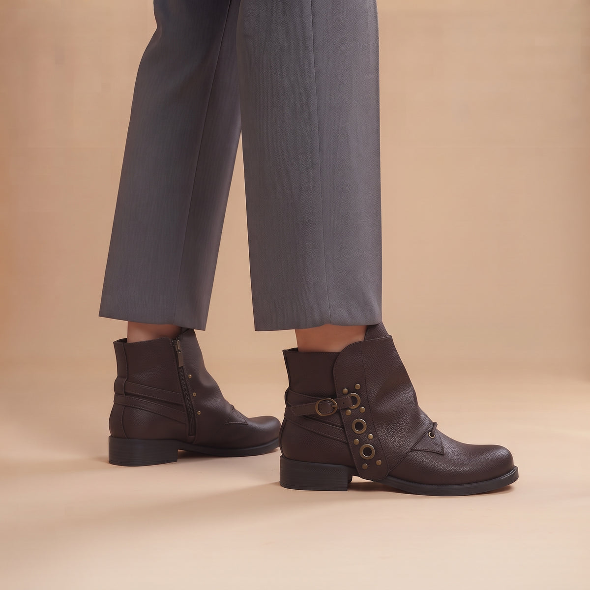 A person wearing dark brown leather ankle boots with decorative metal eyelets and side zippers, paired with gray trousers, against a warm beige studio background.