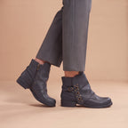 A person wearing dark gray leather ankle boots with side zippers and decorative gold-tone grommets, paired with gray trousers, against a solid beige studio background.