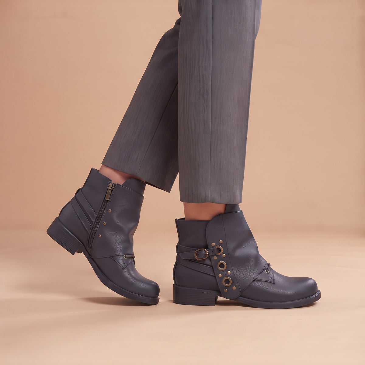 A person wearing dark gray leather ankle boots with side zippers and decorative gold-tone grommets, paired with gray trousers, against a solid beige studio background.