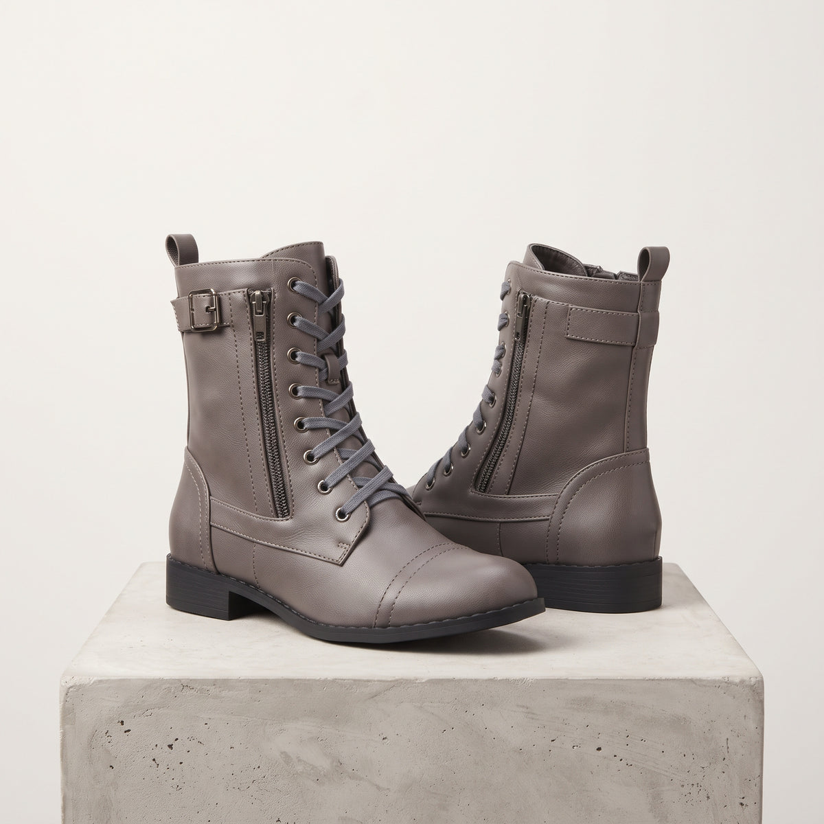 A pair of taupe gray leather lace-up ankle boots with side zippers and buckle details, displayed on a light gray concrete block against a warm off-white background.