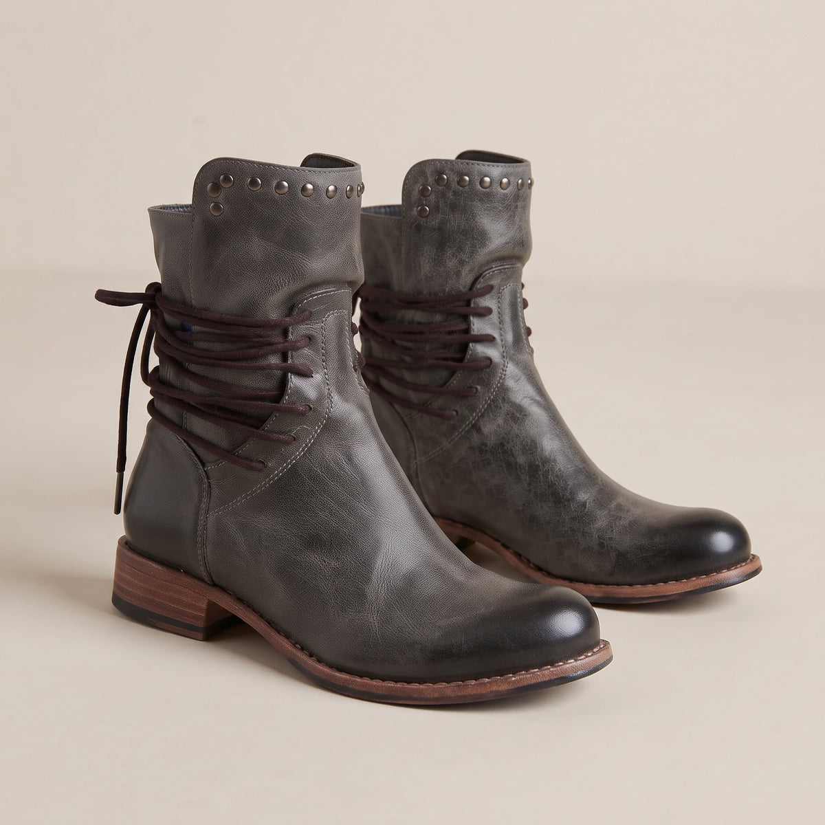 A pair of distressed dark gray leather lace-up ankle boots with silver studs on the collar and a low brown block heel, shown on a neutral beige background.