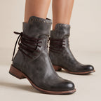 A pair of dark gray distressed leather ankle boots with brown laces, studded detailing near the collar, and low stacked heels, shown on a model against a warm beige background.