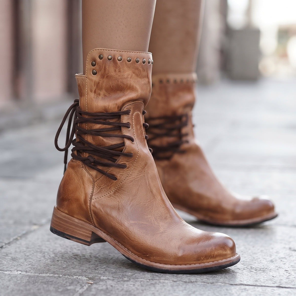 A pair of women's mid-calf brown leather boots with lace-up front, decorative metal studs on the collar, and low block heels, shown worn with tan hosiery on a blurred stone pavement background.