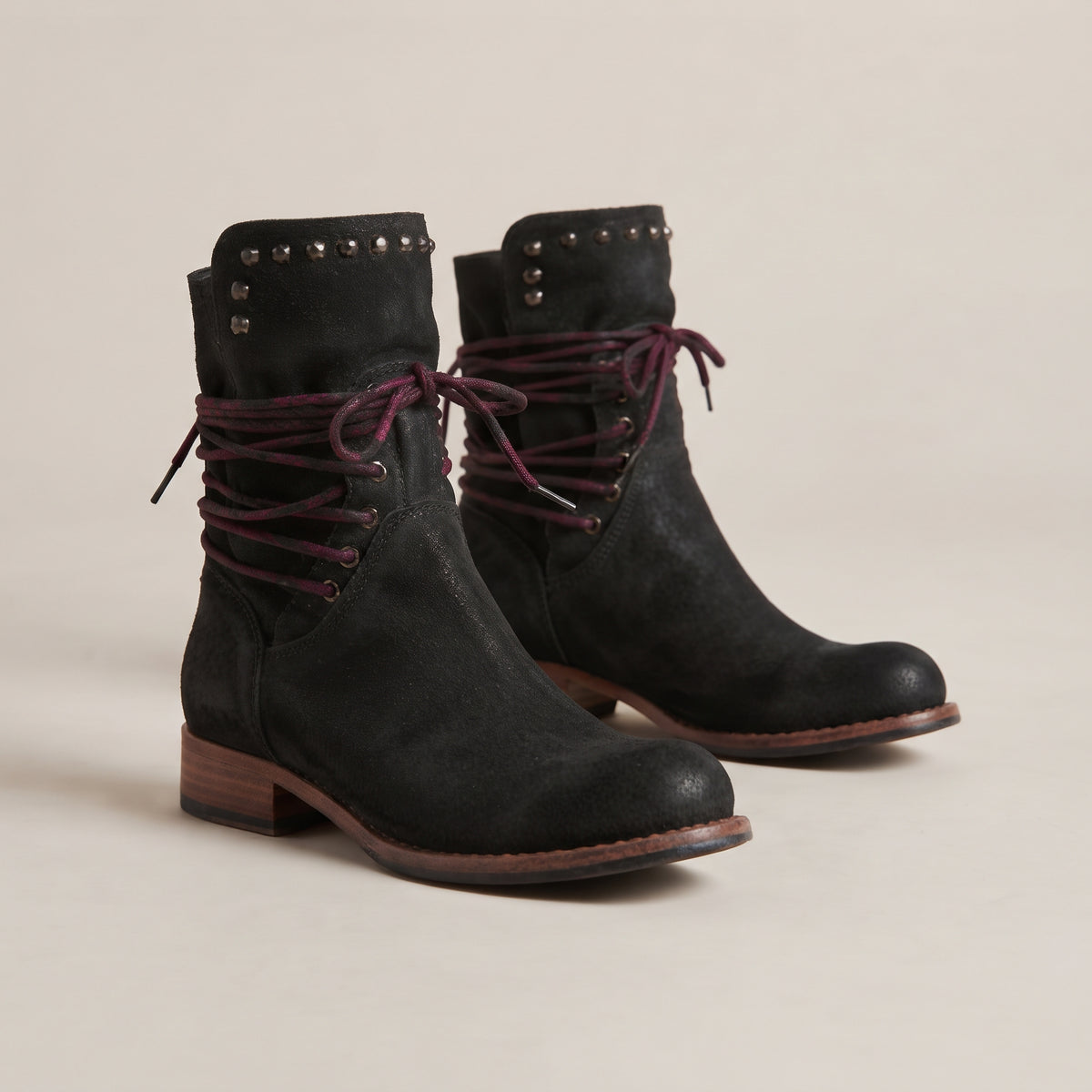 A pair of black suede ankle boots with dark burgundy laces, studded detailing on the collar, and low block heels, photographed on a neutral beige background.