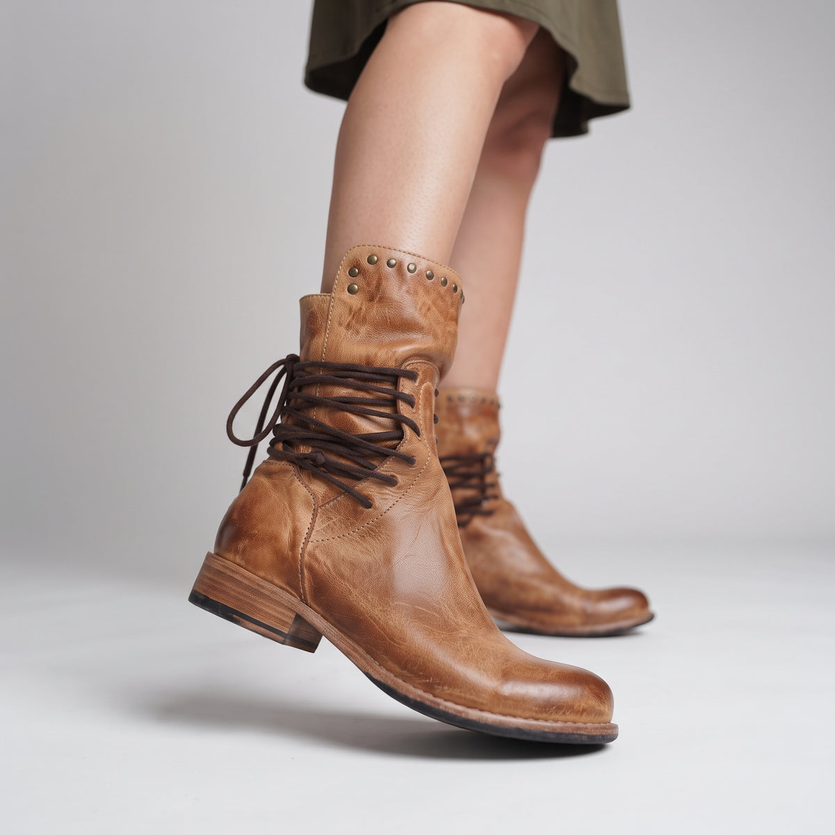 A pair of women's brown leather lace-up ankle boots with a low block heel and decorative metal studs near the collar, shown on a model against a light gray studio background.