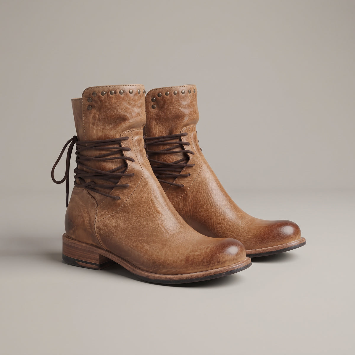 A pair of tan leather lace-up ankle boots with decorative metal studs on the collar and a low stacked heel, photographed on a neutral light gray background.