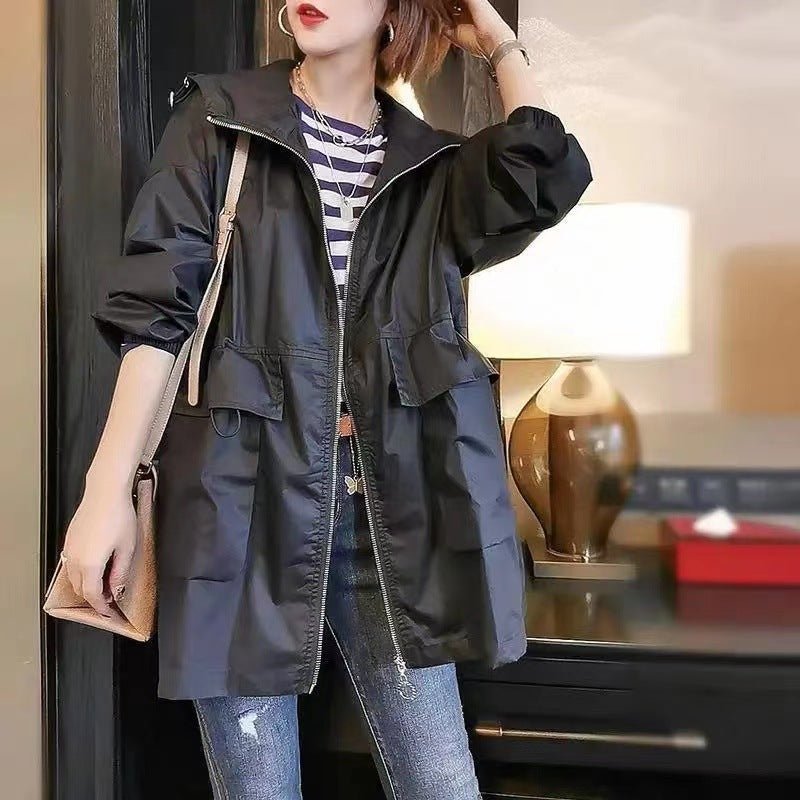 A woman wearing a black hooded utility jacket over a navy and white striped shirt, paired with blue jeans and carrying a beige shoulder bag, standing in an indoor setting with a lamp in the background.