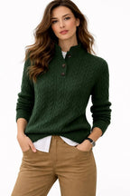 A woman wearing a dark green cable-knit mock-neck sweater with three buttons, styled over a white shirt and tan pants, against a plain white background.