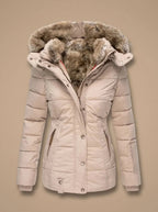 A women's puffer jacket in a light beige or cream color, featuring a plush brown faux-fur trimmed hood and a button-front closure, shown against a solid beige background.