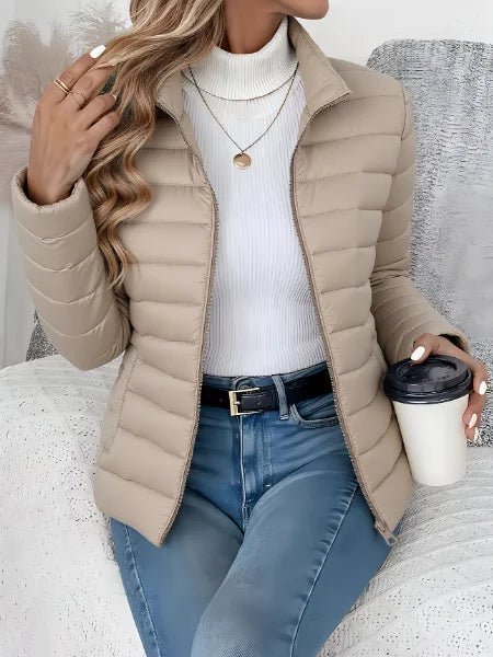 A woman wearing a beige puffer jacket over a white ribbed turtleneck sweater, paired with blue jeans and holding a coffee cup while sitting on a textured white couch.