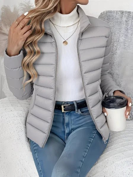 Person wearing a gray puffer jacket, white turtleneck, and blue jeans, holding a coffee cup.