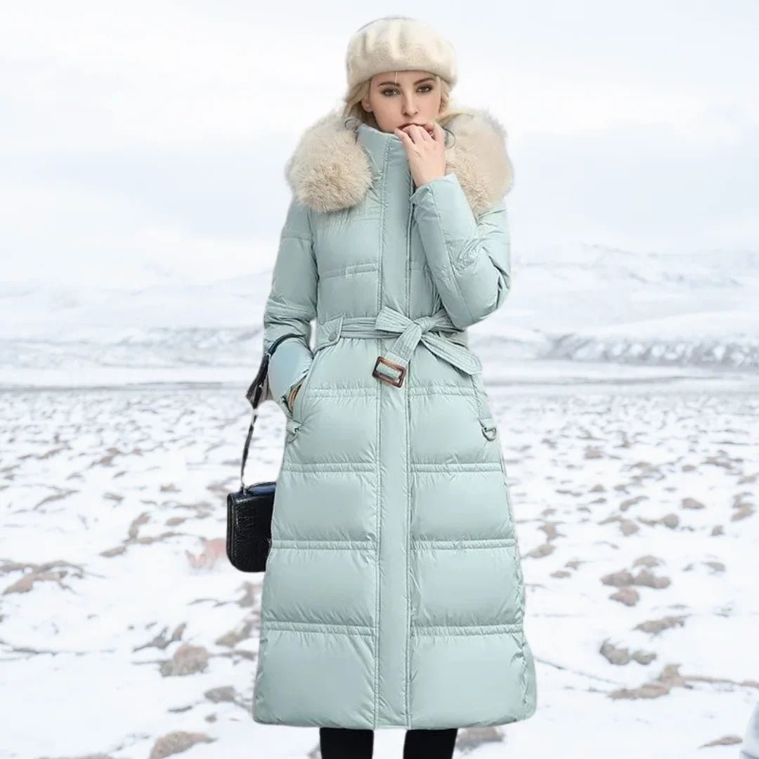 Woman wearing a long light blue puffer coat with a fur-lined hood in a snowy landscape.