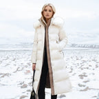 A woman stands in a snowy outdoor landscape wearing a long white puffer coat with a large fluffy hood, over a brown ribbed sweater and black pants.