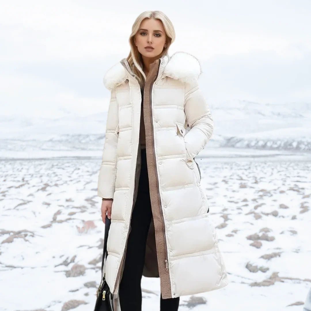 A woman stands in a snowy outdoor landscape wearing a long white puffer coat with a large fluffy hood, over a brown ribbed sweater and black pants.