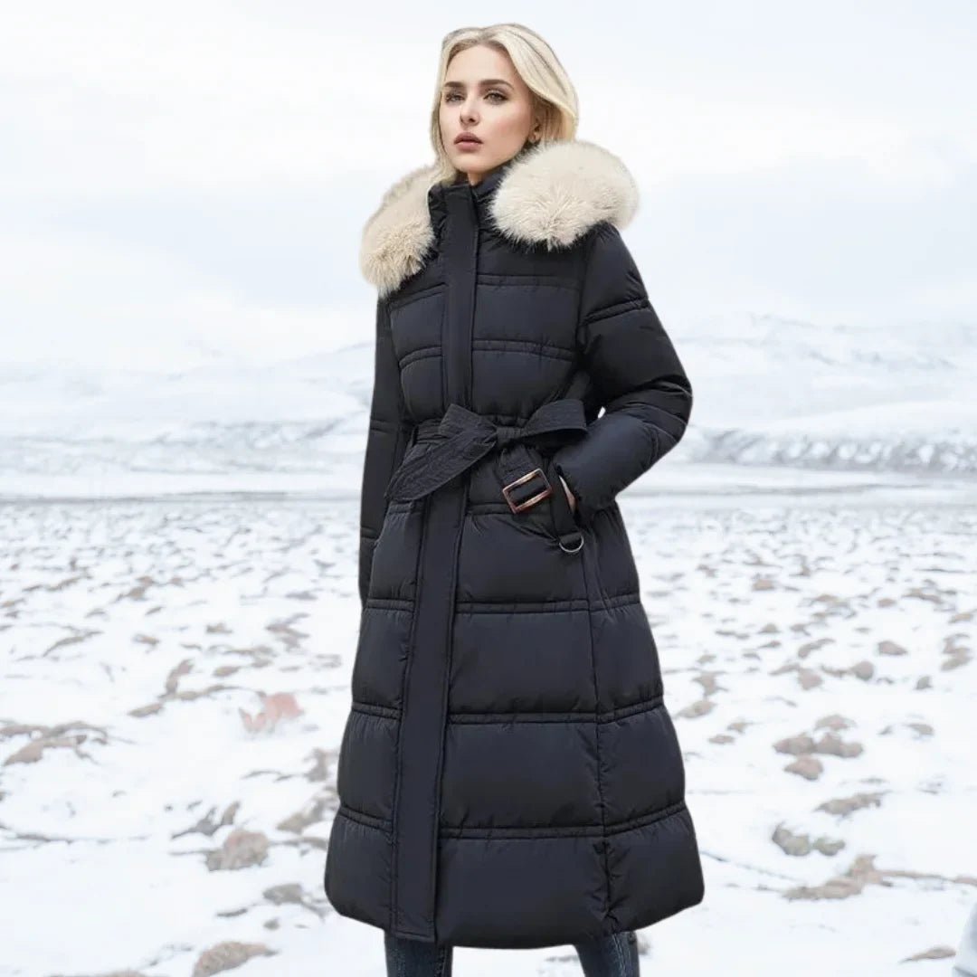 Woman wearing a long black puffer coat with a fur-lined hood in a snowy landscape.