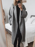 A person taking a mirror selfie in a bedroom, wearing a long, double-breasted gray wool-blend coat over a black outfit.