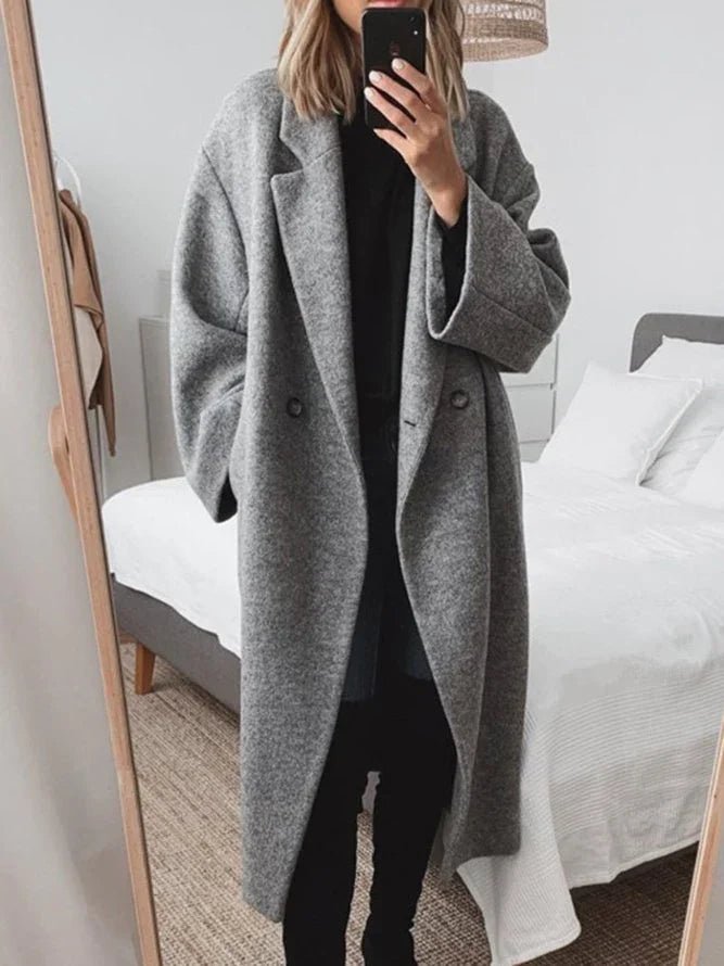 A person taking a mirror selfie in a bedroom, wearing a long, double-breasted gray wool-blend coat over a black outfit.