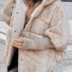 A close-up of a person wearing a beige hooded puffer jacket with faux fur trim and a ribbed knit sleeve, paired with gold rings, against a light stone background.