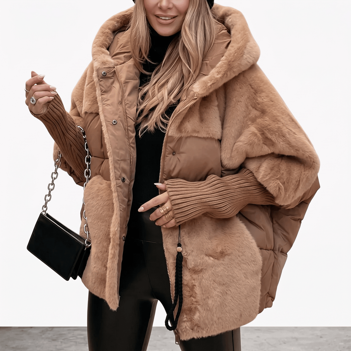 A woman models a chunky, oversized brown faux-fur puffer jacket with a large hood, layered over a black ribbed knit sweater and black pants. She is holding a small black quilted handbag with a silver chain strap against a clean white studio background.