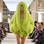 Model wearing a bright green fur coat and dress on a fashion runway.