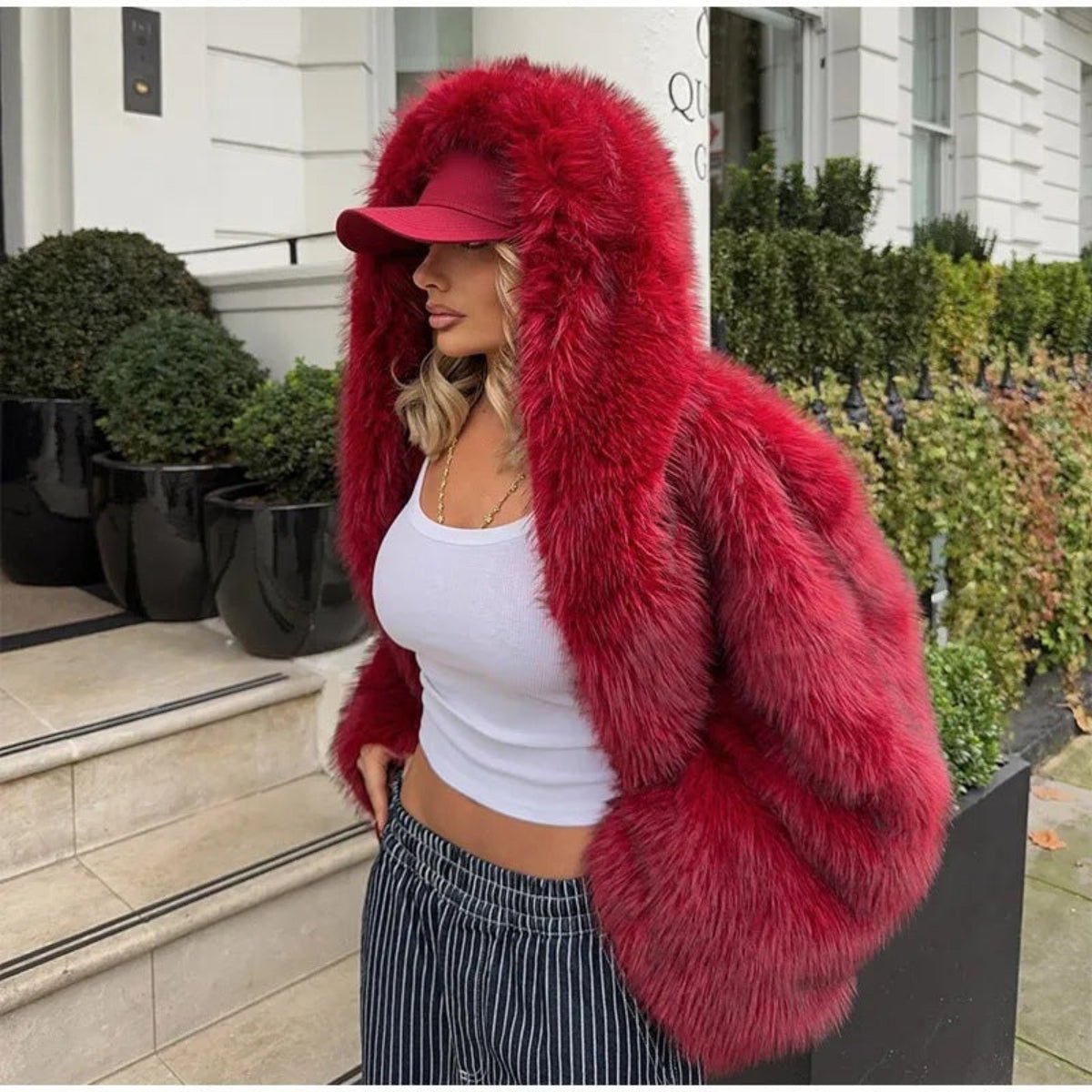 A woman wearing a voluminous bright red faux fur hooded jacket with a matching red baseball cap, posing outdoors in front of a white building with green hedges.