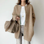 A woman models an oversized, fuzzy beige knit cardigan over a white t-shirt and light gray jeans, carrying a matching beige leather handbag against a plain light gray background.