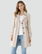 Woman wearing a beige trench coat over a white shirt and blue jeans on a light gray background