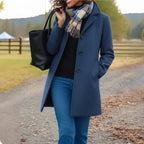 Woman wearing a blue coat with a scarf, holding a black handbag, standing on a road with a scenic background.