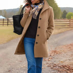 Woman wearing a beige coat and blue jeans, holding a black bag with a scenic background.