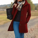 A woman wearing a long burgundy wool-blend coat with a high collar and dark buttons, styled with a beige plaid scarf, a black top, blue jeans, and carrying a black tote bag, standing on a country path.