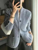A person wearing a light gray textured knit cardigan with a stand collar and front buttons, paired with a white top and dark skirt. They are holding a black smartphone to take a mirror selfie, with a white handbag and gold jewelry visible.